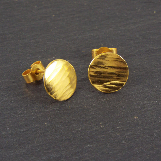 A pair of disc-shaped hammered silver earrings, 10mm across, plated with yellow gold with a satin-matte finish. The hammered texture of the studs is strongly horizontal.
