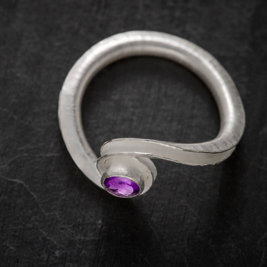 A silver birthstone ring handmade from recycled sterling silver, with a gemstone cradled between two curling ends. Shown here is the amethyst variant for February, from above and the front.