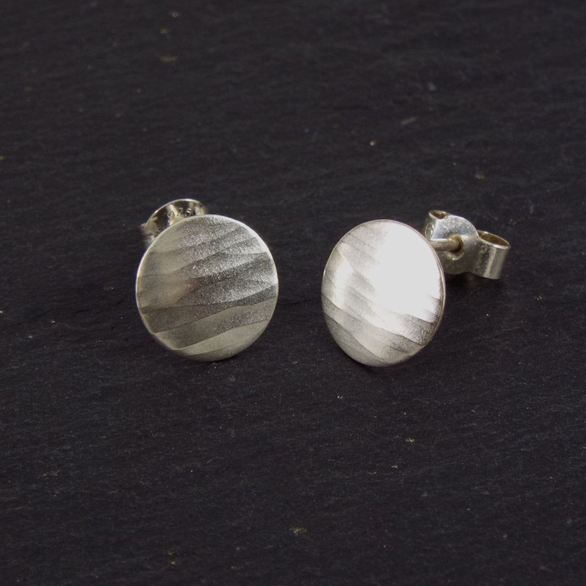 A pair of disc-shaped hammered silver earrings, 10mm across, with a satin-matte finish. The hammered texture of the studs is strongly horizontal.