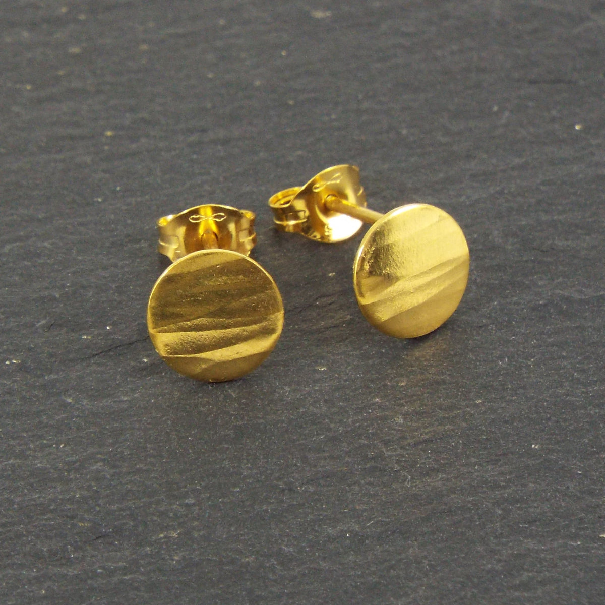 A pair of disc-shaped hammered silver earrings, 7.5mm across, plated with yellow gold with a satin-matte finish. The hammered texture of the studs is strongly horizontal.