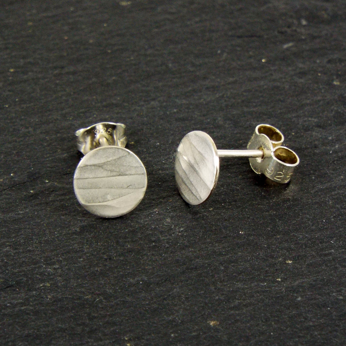 A pair of disc-shaped hammered silver earrings, 7.5mm across, with a satin-matte finish. The hammered texture of the studs is strongly horizontal.