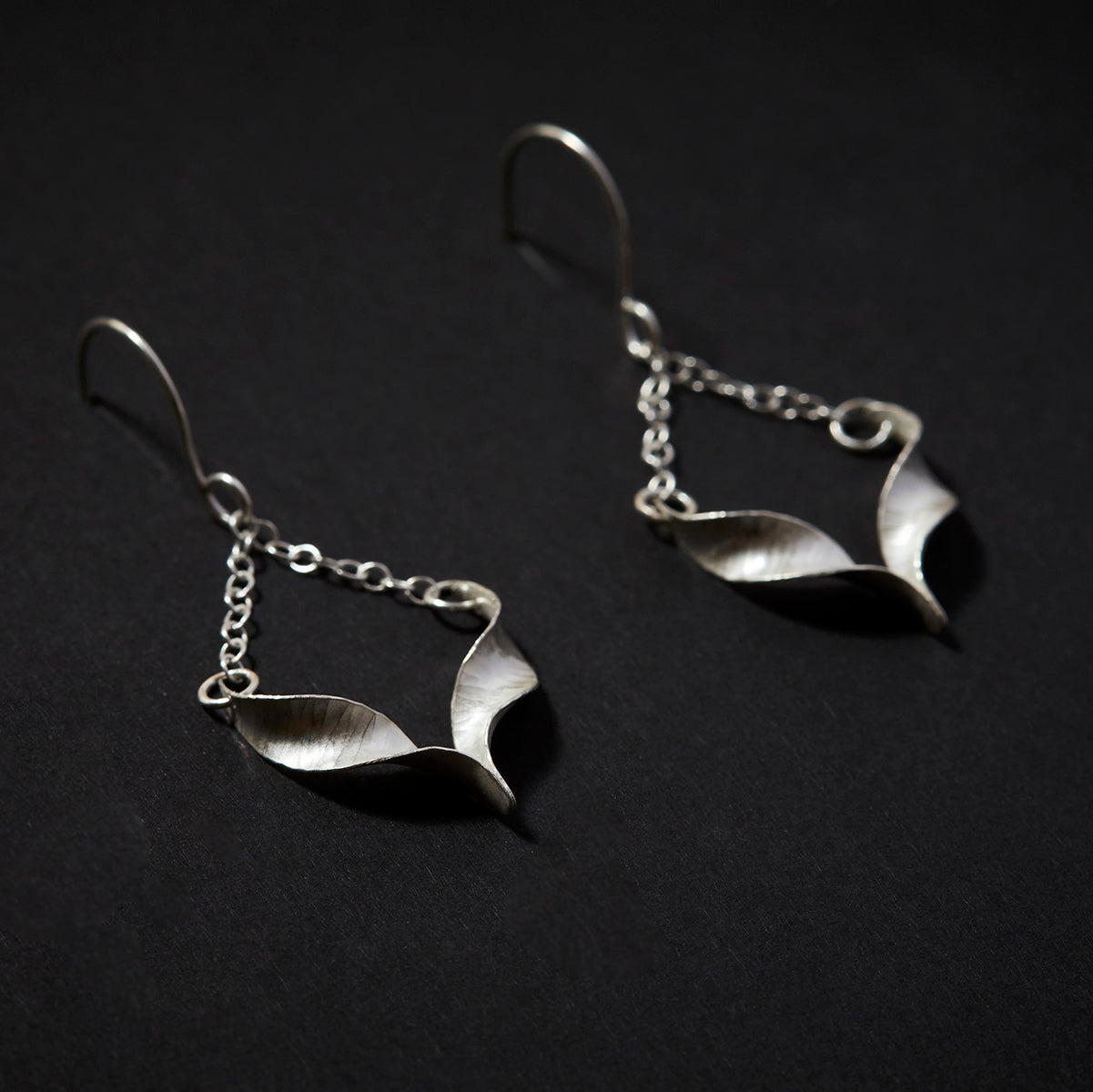 A pair of sculptural silver bird earrings. Each is composed of two twists of hammered silver joined at the central point and hanging from a handmade hook by means of delicate trace chain. They have a hammered texture, non-reflective finish and burnished edges. Shown lying at an angle.