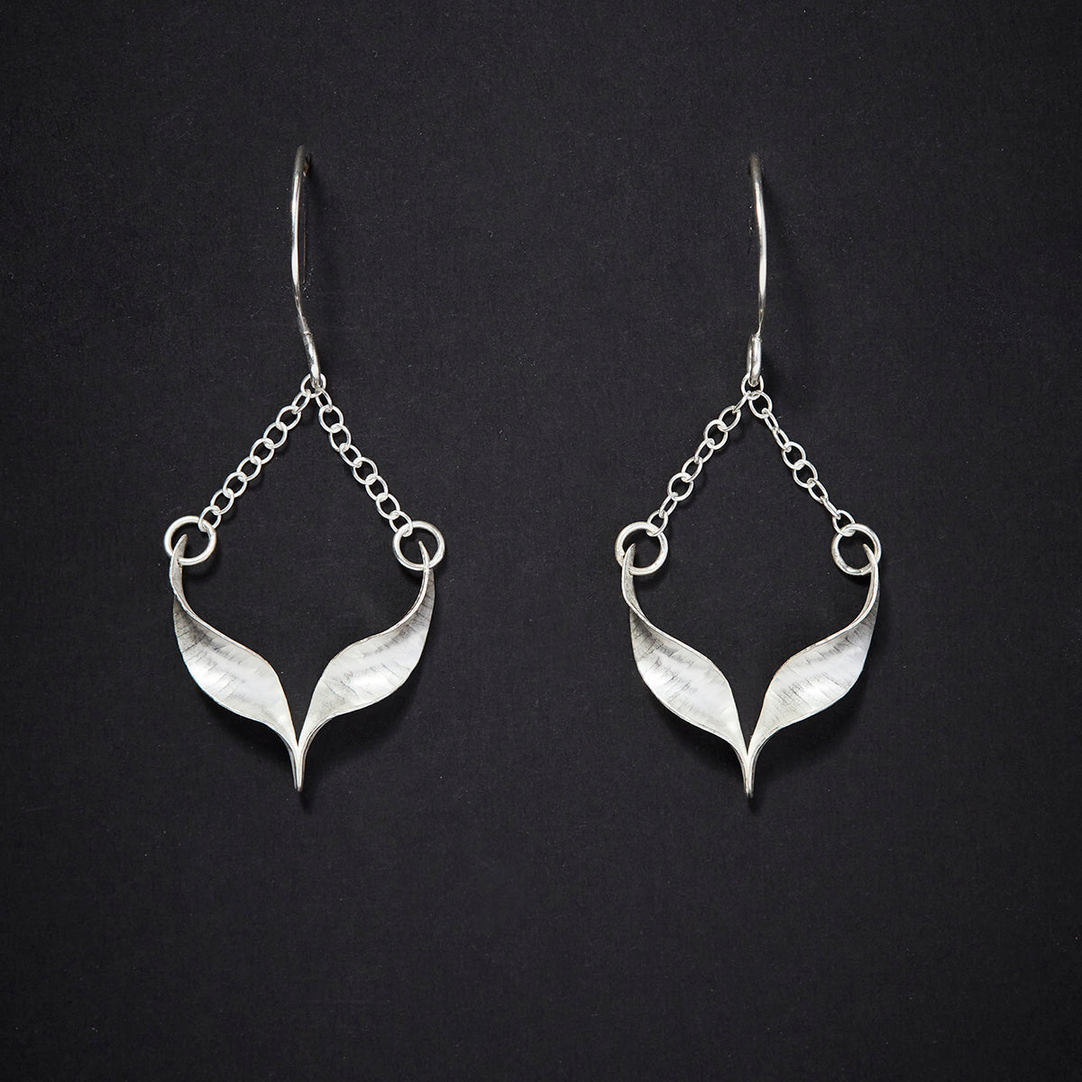 A pair of sculptural silver bird earrings. Each is composed of two twists of hammered silver joined at the central point and hanging from a handmade hook by means of delicate trace chain. They have a hammered texture, non-reflective finish and burnished edges. Shown from the front.