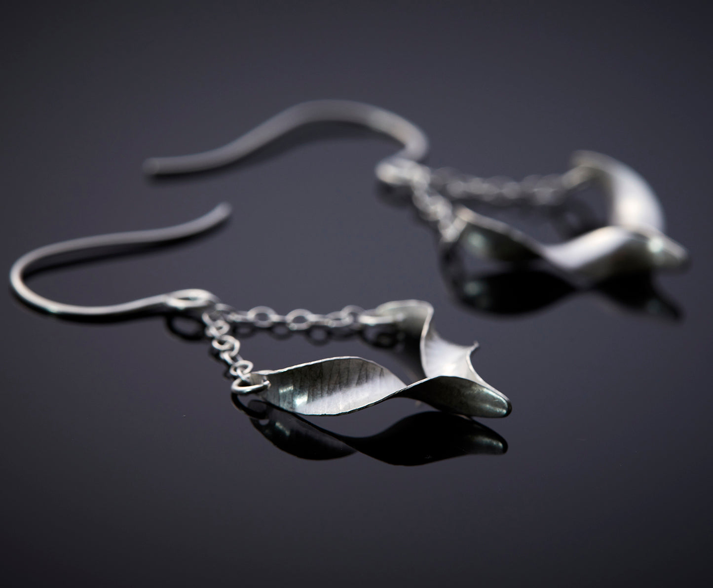 A pair of sculptural silver bird earrings. Each is composed of two twists of hammered silver joined at the central point and hanging from a handmade hook by means of delicate trace chain. They have a hammered texture, non-reflective finish and burnished edges. Shown lying at an angle so that the twist is emphasised and the central join displayed.