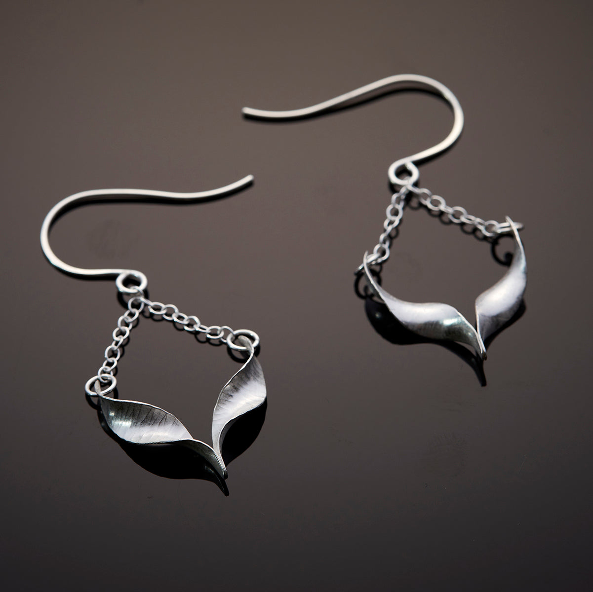 A pair of sculptural silver bird earrings. Each is composed of two twists of hammered silver joined at the central point and hanging from a handmade hook by means of delicate trace chain. They have a hammered texture, non-reflective finish and burnished edges. The front of one is shown with its hammered texture; the smooth back of the other is on display.