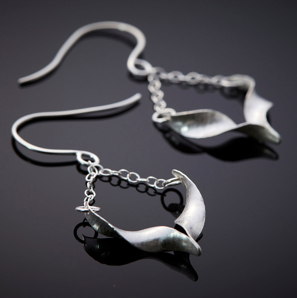 A pair of sculptural silver bird earrings. Each is composed of two twists of hammered silver joined at the central point and hanging from a handmade hook by means of delicate trace chain. They have a hammered texture, non-reflective finish and burnished edges. The back of one of the earrings is shown in the foreground, from an angle which shows the central join.