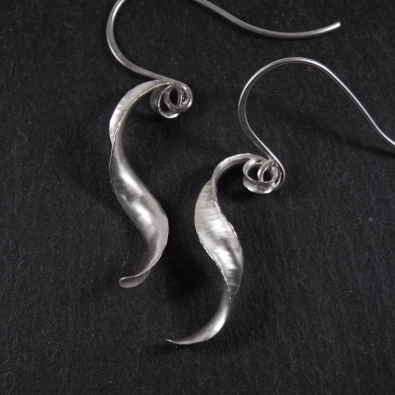 A pair of s-shaped earrings made from recycled sterling silver by the technique of anticlastic raising. The two earrings are mirror images of one another and have a curling, twisting shape. The right hand one is shown from the back and the other from the front.