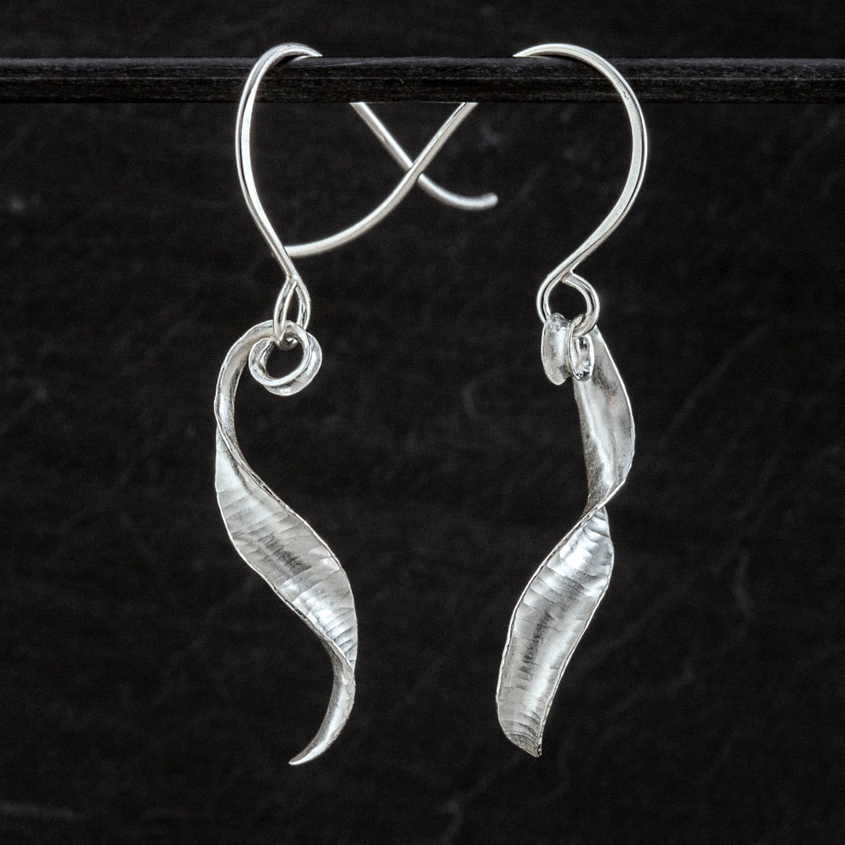 A pair of s-shaped earrings made from recycled sterling silver by the technique of anticlastic raising. The two earrings are mirror images of one another and have a curling, twisting shape. The right hand one is shown from the front and the other from the side.