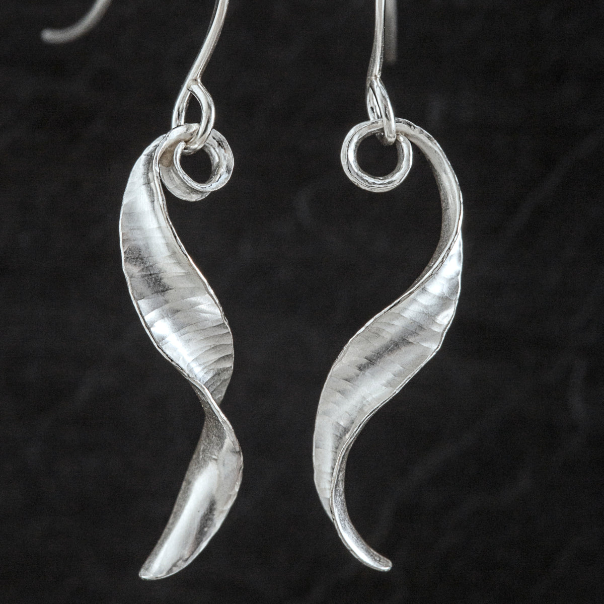 A pair of s-shaped earrings made from recycled sterling silver by the technique of anticlastic raising. The two earrings are mirror images of one another and have a curling, twisting shape. The right hand one is shown from the side and the other from the front.