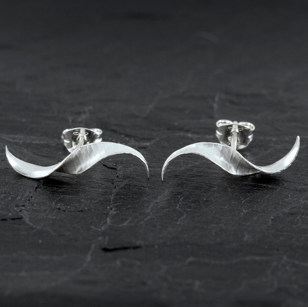 A pair of simple mirror-image silver twist earrings. They each have a stretched-S shape, a hammered texture, non-shiny finish and glittering edges. They are shown on a slate background.