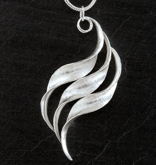A silver wave necklace, handmade, composed of three curled and twisted units, the top one ending in a loop for the chain. The three units are arranged so that they form a progression.