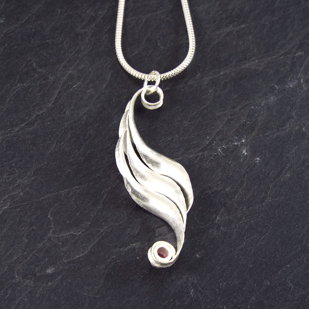 A silver wave necklace, handmade, composed of three curled and twisted units, the top one ending in a loop for the chain and the bottom one curled around a 3mm rhodolite garnet, faceted, magenta. The three units are arranged so that they form a progression. This is a back view.