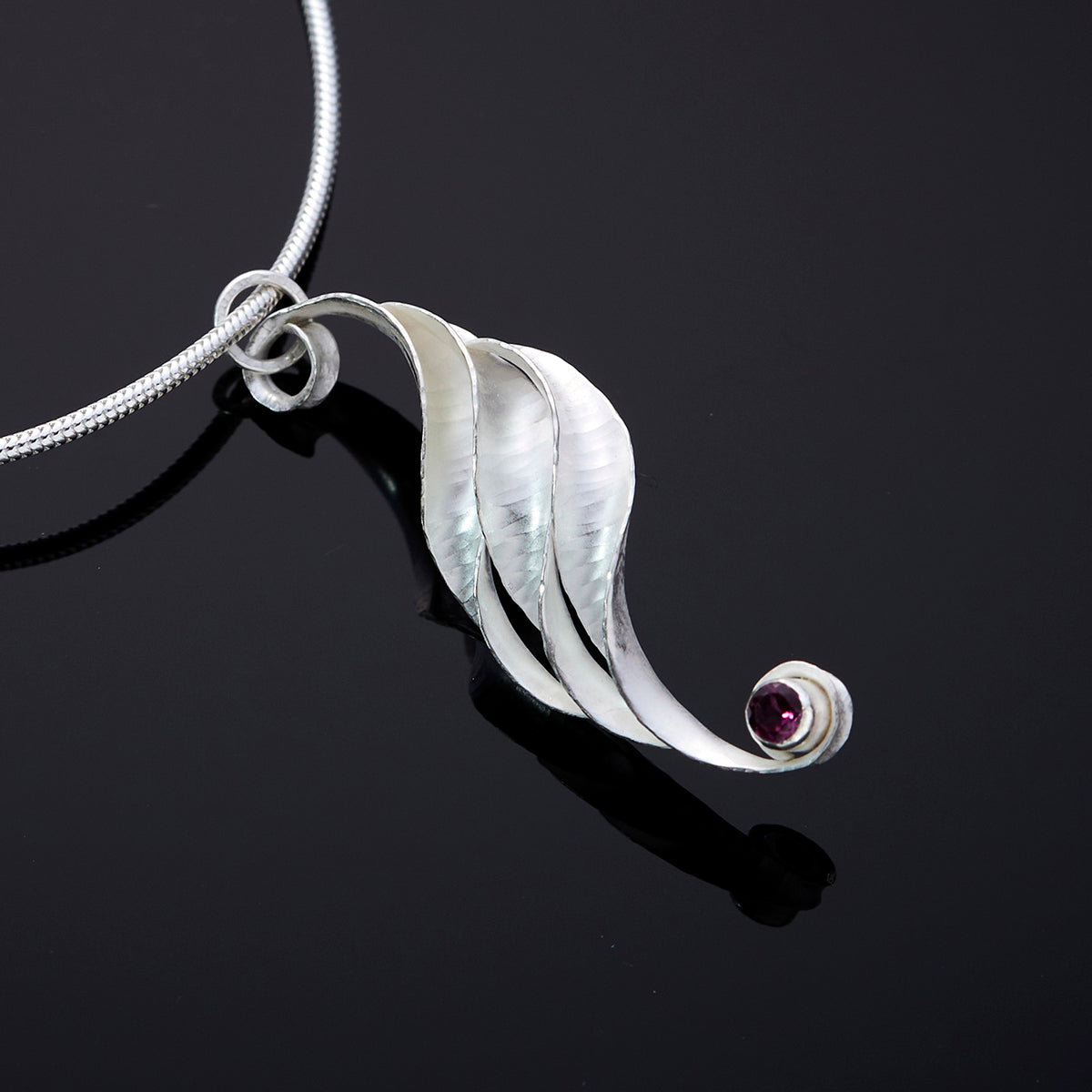 A silver wave necklace, handmade, composed of three curled and twisted units, the top one ending in a loop for the chain and the bottom one curled around a 3mm rhodolite garnet, faceted, magenta. The three units are arranged so that they form a progression.