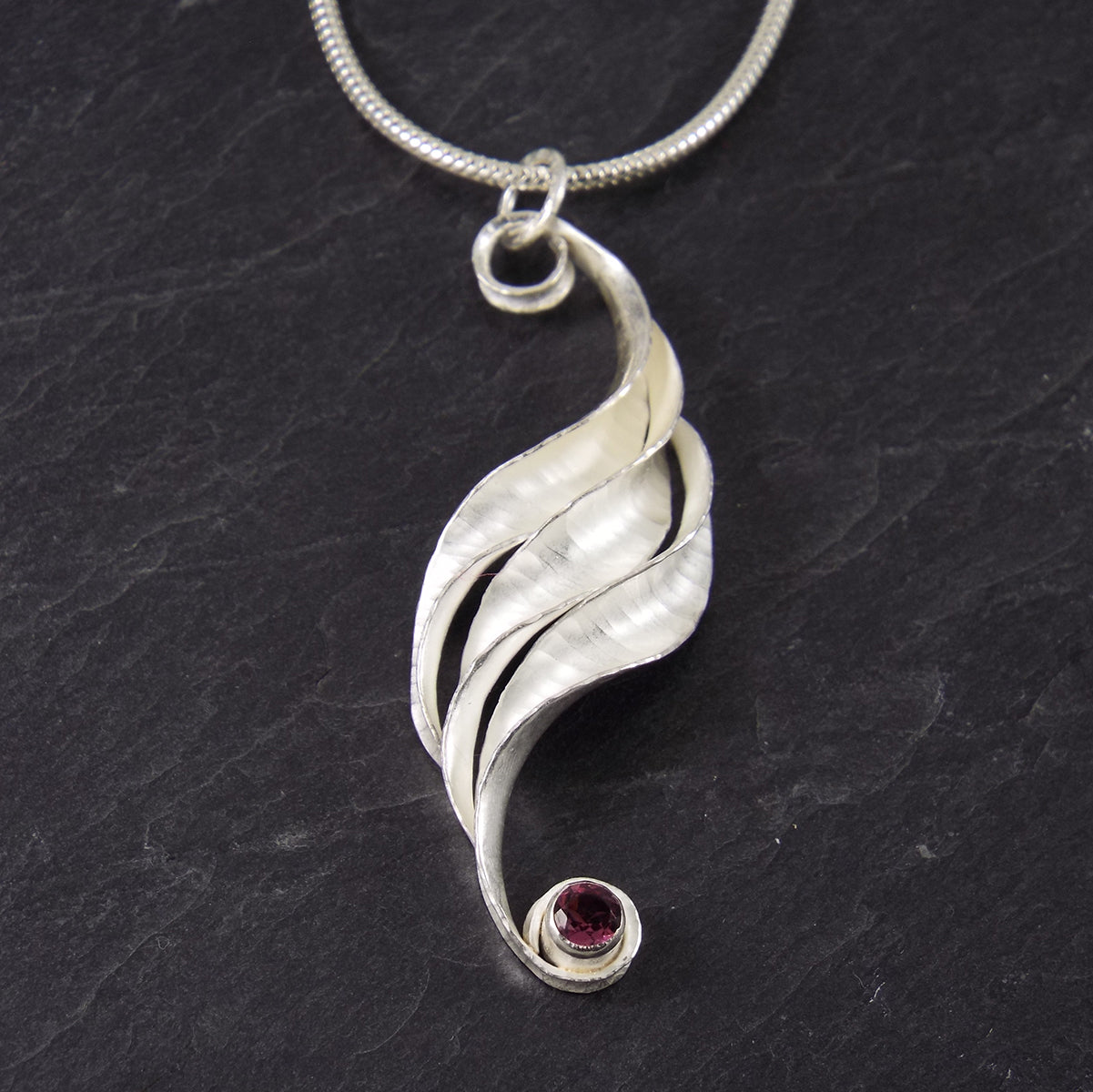 A silver wave necklace, handmade, composed of three curled and twisted units, the top one ending in a loop for the chain and the bottom one curled around a 3mm rhodolite garnet, faceted, magenta. The three units are arranged so that they form a progression.