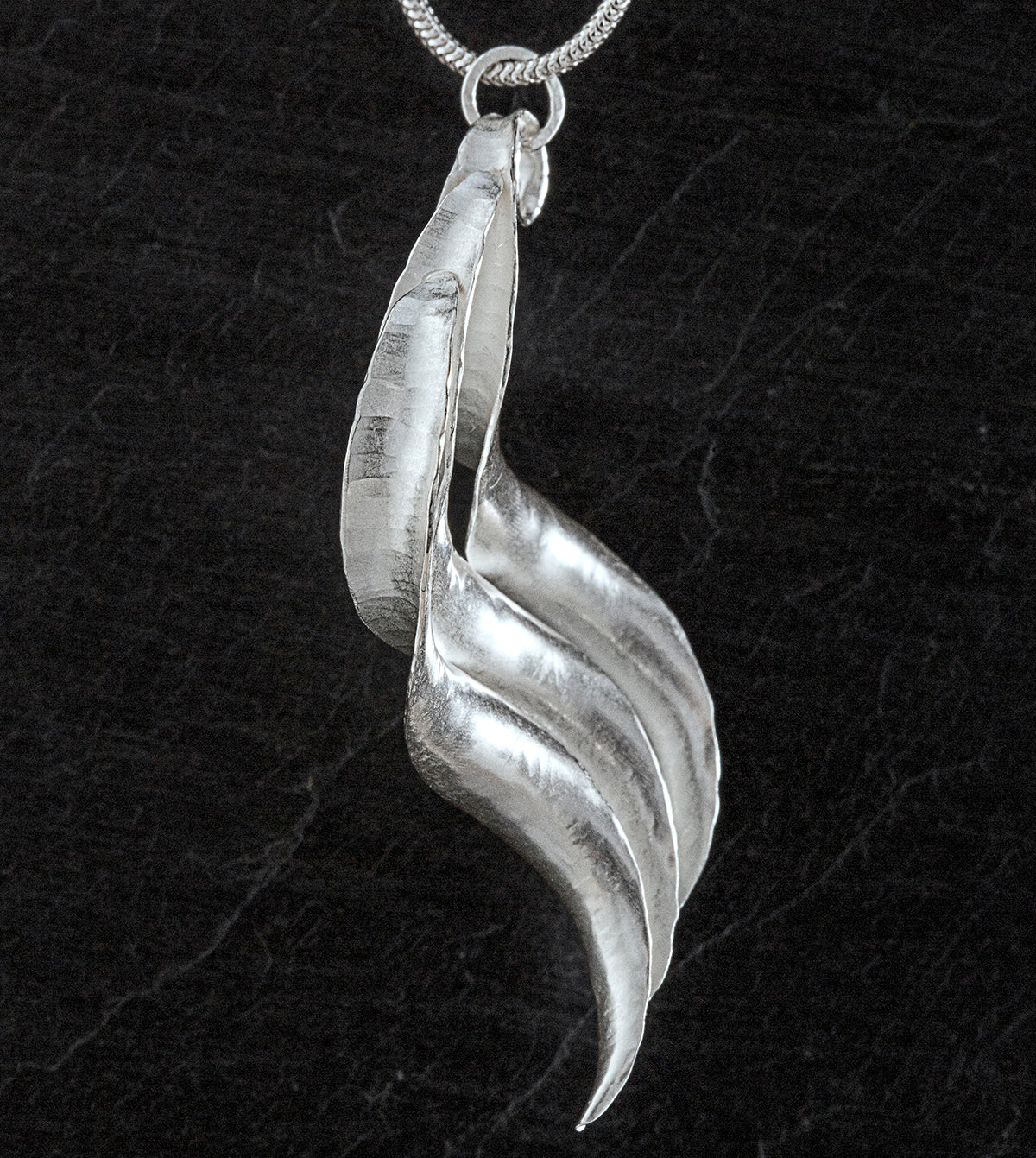 A silver pendant, handmade, composed of three curled and twisted units, the top one ending in a loop for the chain. The three units are arranged so that they form a progression. Shown from the side.