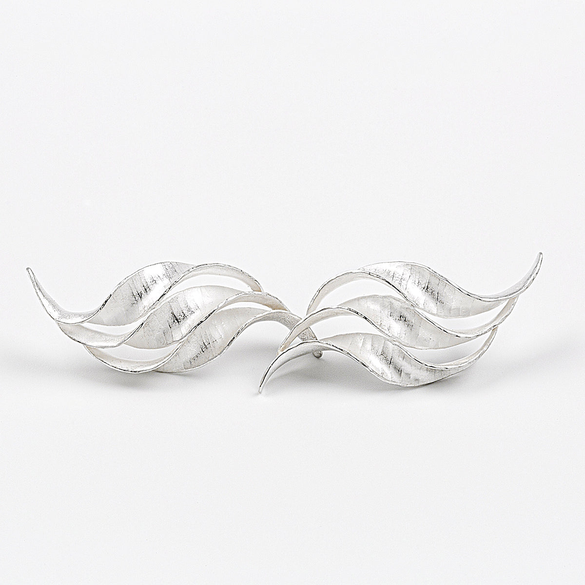 A pair of silver wedding earrings in the form of a triple wave of parallel curling, twisting S-shapes. They have a hammered texture, non-reflective surface and burnished edges. 