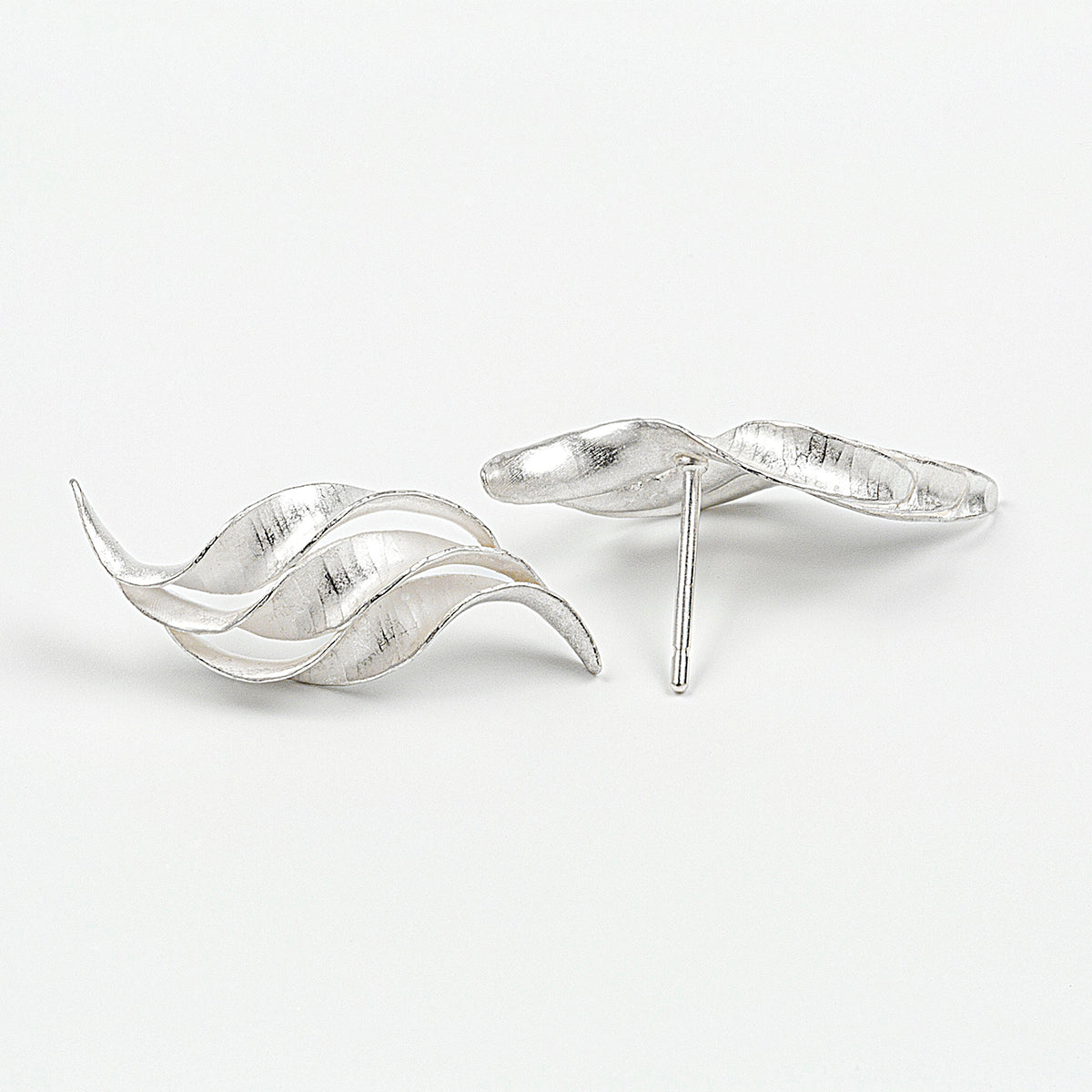 A pair of silver wedding earrings in the form of a triple wave of parallel curling, twisting S-shapes. They have a hammered texture, non-reflective surface and burnished edges. This view shows one earring from the front and the other showing how the pin attaches at the back.