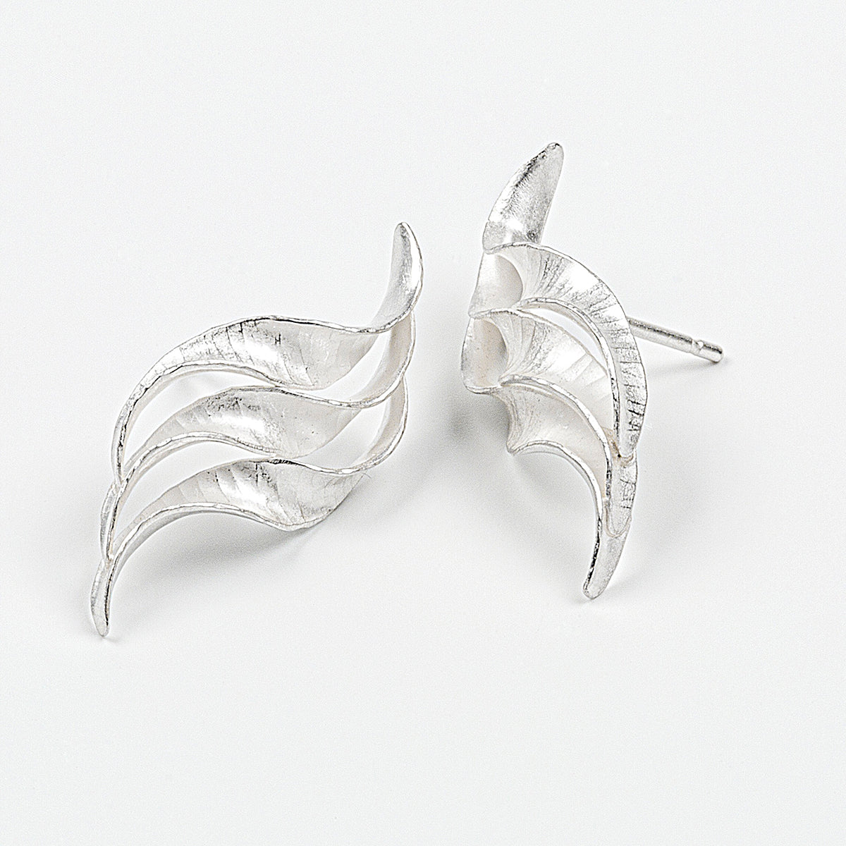 A pair of silver wedding earrings in the form of a triple wave of parallel curling, twisting S-shapes. They have a hammered texture, non-reflective surface and burnished edges.  This view shows the two studs turned slightly to face one another.