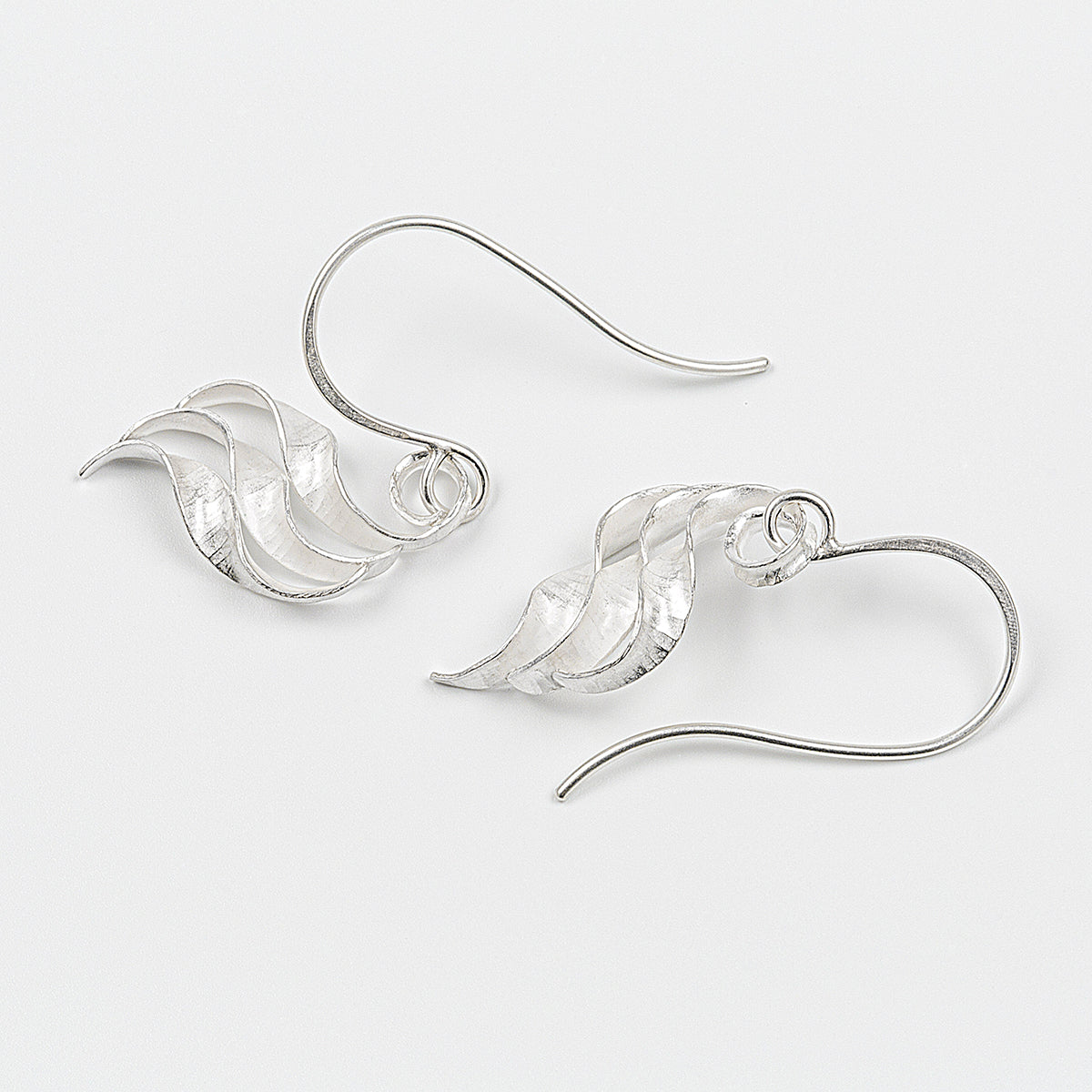 A pair of silver wing earrings made from recycled sterling silver by the process of anticlastic raising. These drop earrings have a hammered texture and non-reflective finish. Each consists of three s-shaped twisted units nested together. A different view of the smaller earrings.