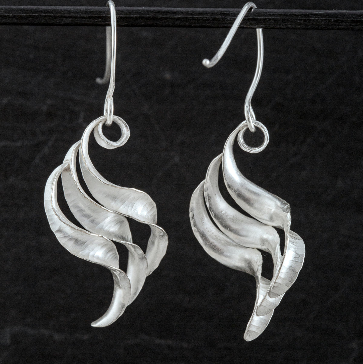 A pair of silver wing earrings made from recycled sterling silver by the process of anticlastic raising. These drop earrings have a hammered texture and non-reflective finish. Each consists of three s-shaped twisted units nested together. Here one is shown fronm the front and one from the back.