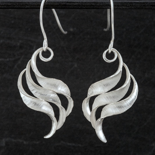 A pair of silver wing earrings made from recycled sterling silver by the process of anticlastic raising. These drop earrings have a hammered texture and non-reflective finish. Each consists of three s-shaped twisted units nested together. This image shows both from the front.