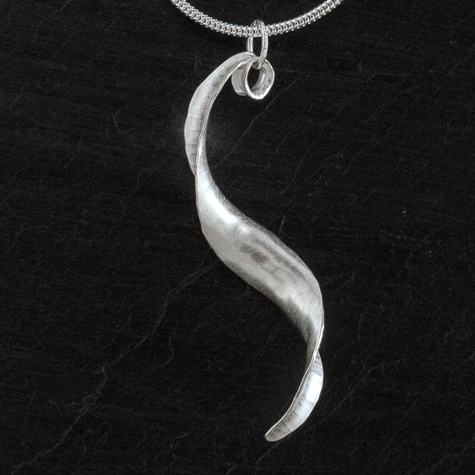 Handmade twisted silver necklace made from recycled silver with hammered texture, burnished edges and satin-matte finish, hanging from a sterling silver snake chain. It has the shape of a reversed s and an itegral loop by which it hangs from the chain. Back view.
