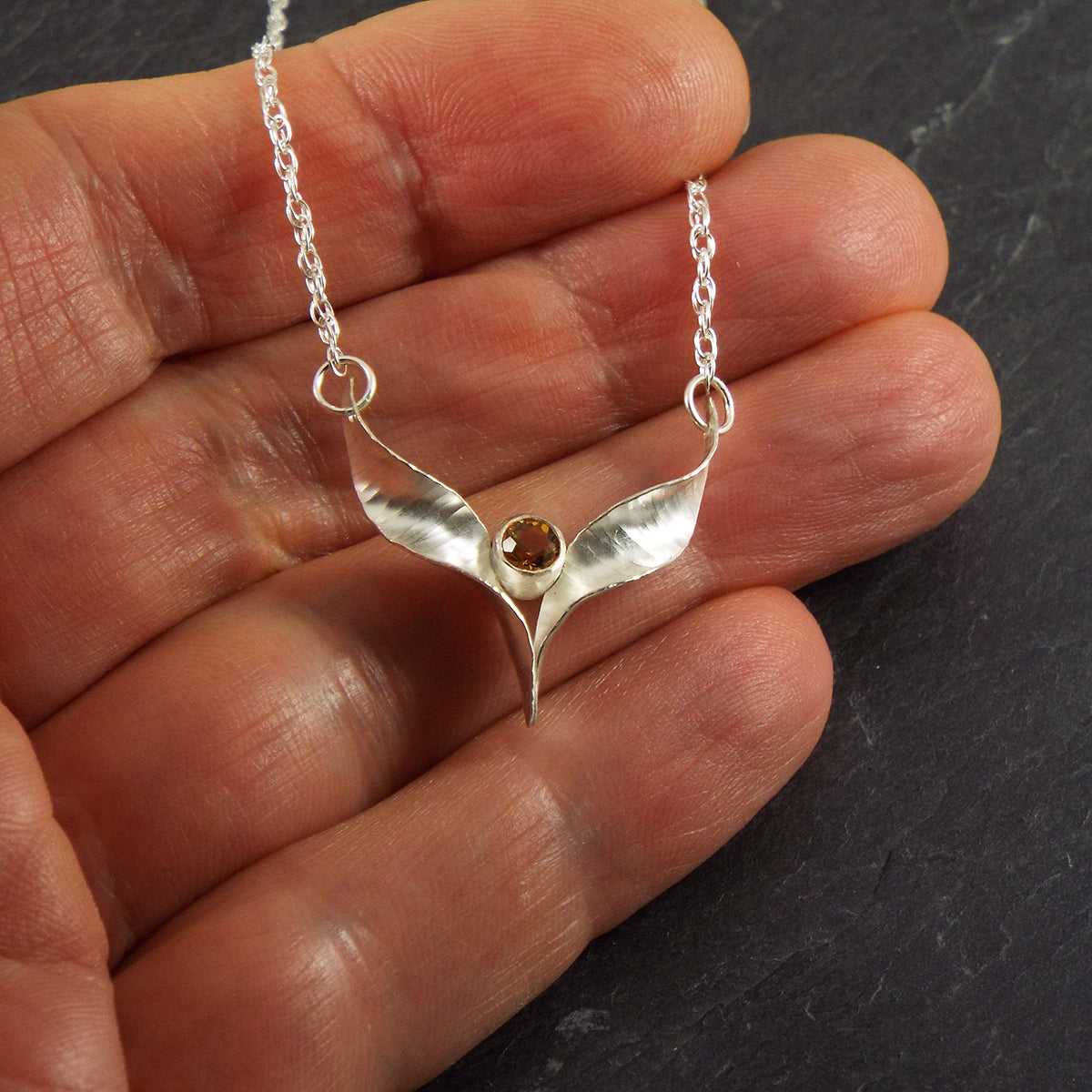 A silver bird necklace made from two mirror-image twisted and curled units soldered at the lower point and attached to a silver rope chain by a silver jump ring at each outer point. This one is set with a yellow citrine. It is shown held in the hand to give an idea of the scale.
