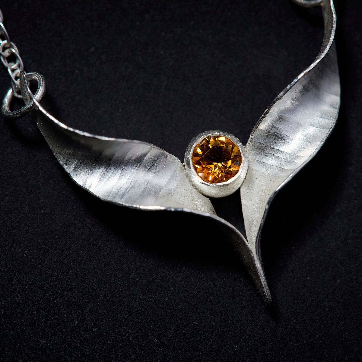 A silver bird necklace made from two mirror-image twisted and curled units soldered at the lower point and attached to a silver rope chain by a silver jump ring at each outer point. A sparkling yellow citrine is set in a length of silver tube which sits above the lower point where the two units meet.