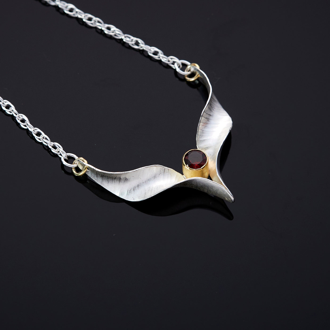 A silver bird necklace made from two mirror-image twisted and curled units. They are  soldered at the lower point and attached to a silver rope chain by an 18 carat gold  jump ring at each outer point. A sparkling red garnet is set in a length of 18 carat gold tube which sits above the lower point where the two units meet.