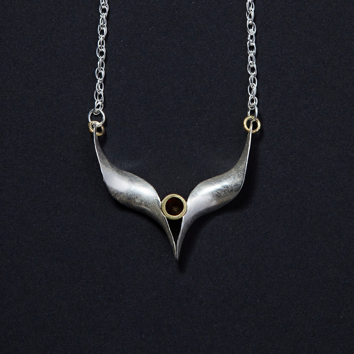 A silver bird necklace made from two mirror-image twisted and curled units. They are soldered at the lower point and attached to a silver rope chain by an 18 carat gold jump ring at each outer point. A sparkling red garnet is set in a length of 18 carat gold tube which sits above the lower point where the two units meet. View from the back.