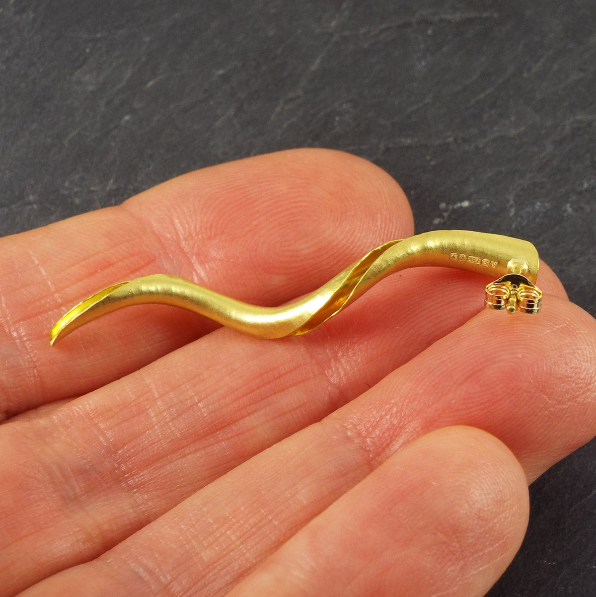 A pair of mirror image gold spiral stud earrings, each in the form of a tapered spiralling tube with one side open to show the inner surface. They have a satin matte finish and hammered texture. Shown here in the hand to demonstrate scale and to show the hallmark.