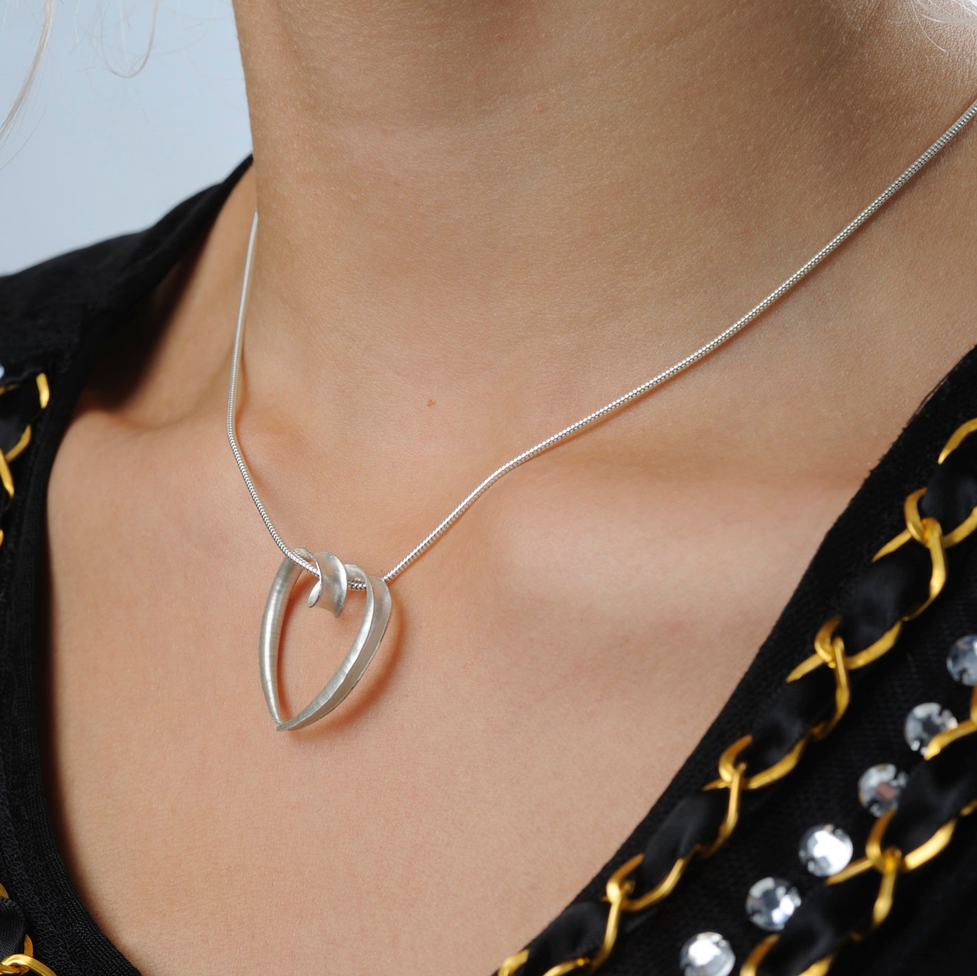 A large silver heart pendant made from a single piece of metal, formed into a sort of tube with the outer side open. It is formed into a loop at the top which the chain passes through. Shown here on a model.