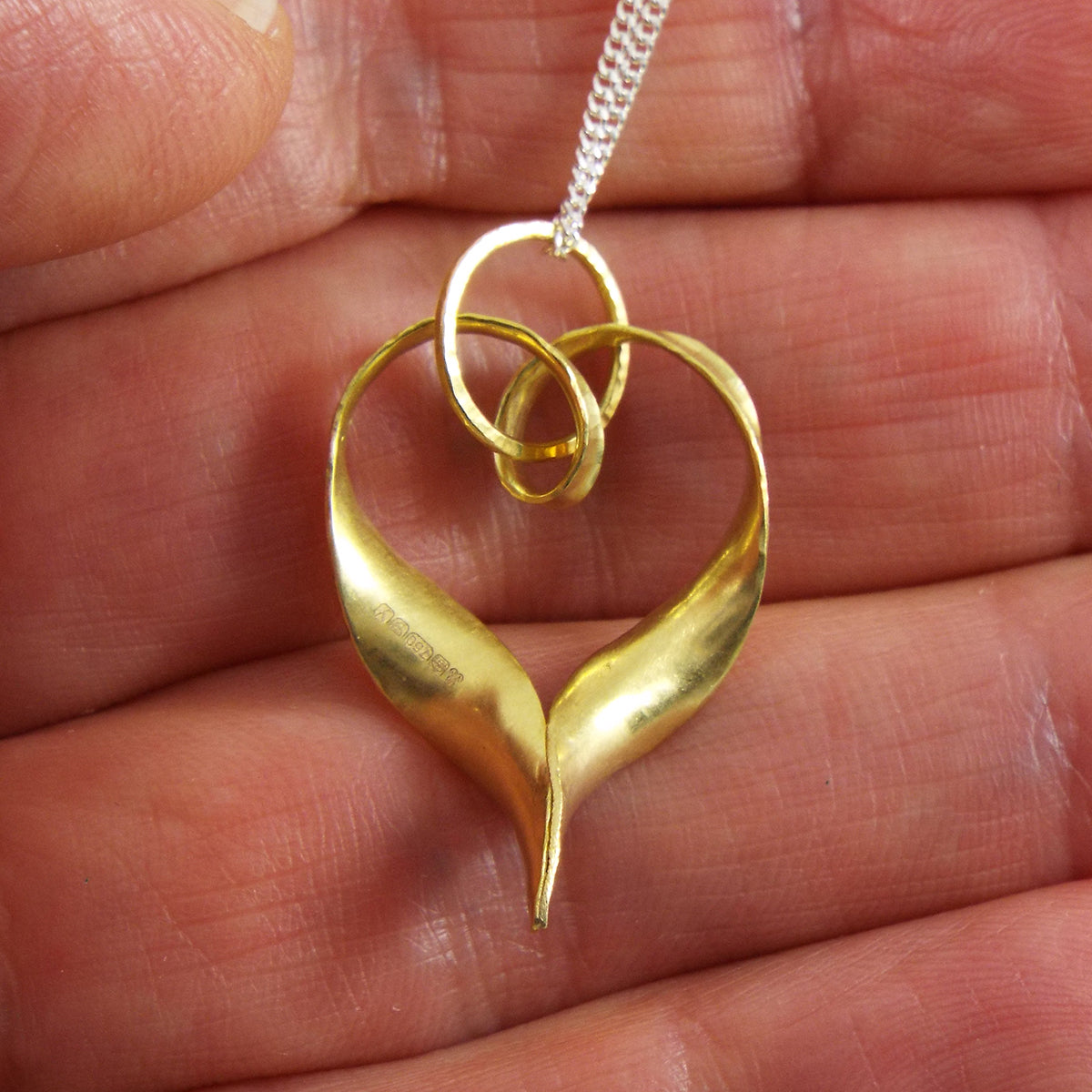 A recycled 18 carat gold heart pendant made from a ribbon of metal, hammered into a continuous heart shape, with a loop at the top for the chain to pass through and elegant twisted ends which join in a point at the bottom. This is a back view, showing the hallmark and the scale relative to the hand.