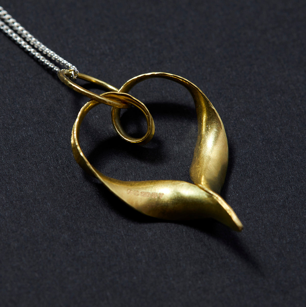 A recycled 18 carat gold heart pendant made from a ribbon of metal, hammered into a continuous heart shape, with a loop at the top for the chain to pass through and elegant twisted ends which join in a point at the bottom. This is a back view, showing the hallmark.