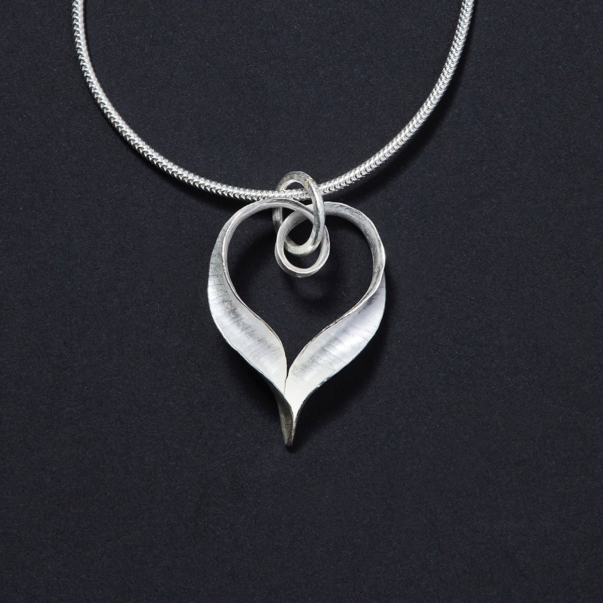 A recycled silver heart pendant made from a ribbon of metal, hammered into a continuous heart shape, with a loop at the top for the chain to pass through and elegant twisted ends which join in a point at the bottom.
