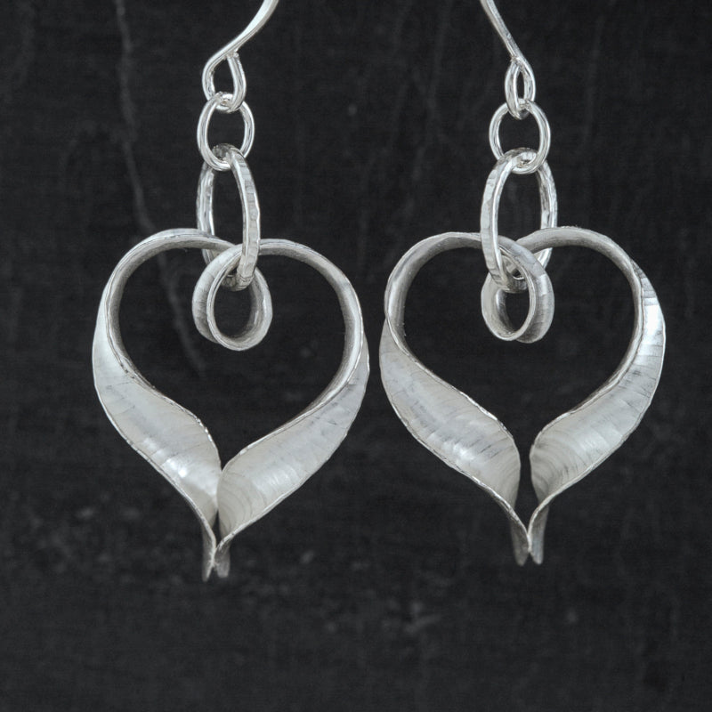 A pair of curving, twisting silver heart drop earrings, each formed from a single ribbon of silver, with a loop at the top for the jump ring and joined at the bottom, pointed end. They have a hammered texture and non-reflective finish.