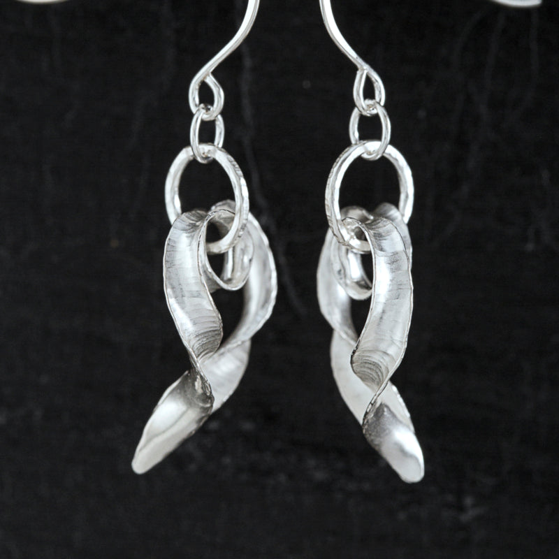 A pair of silver heart drop earrings, curling and twisting, viewed from the side, showing the hammered texture and burnished edges.