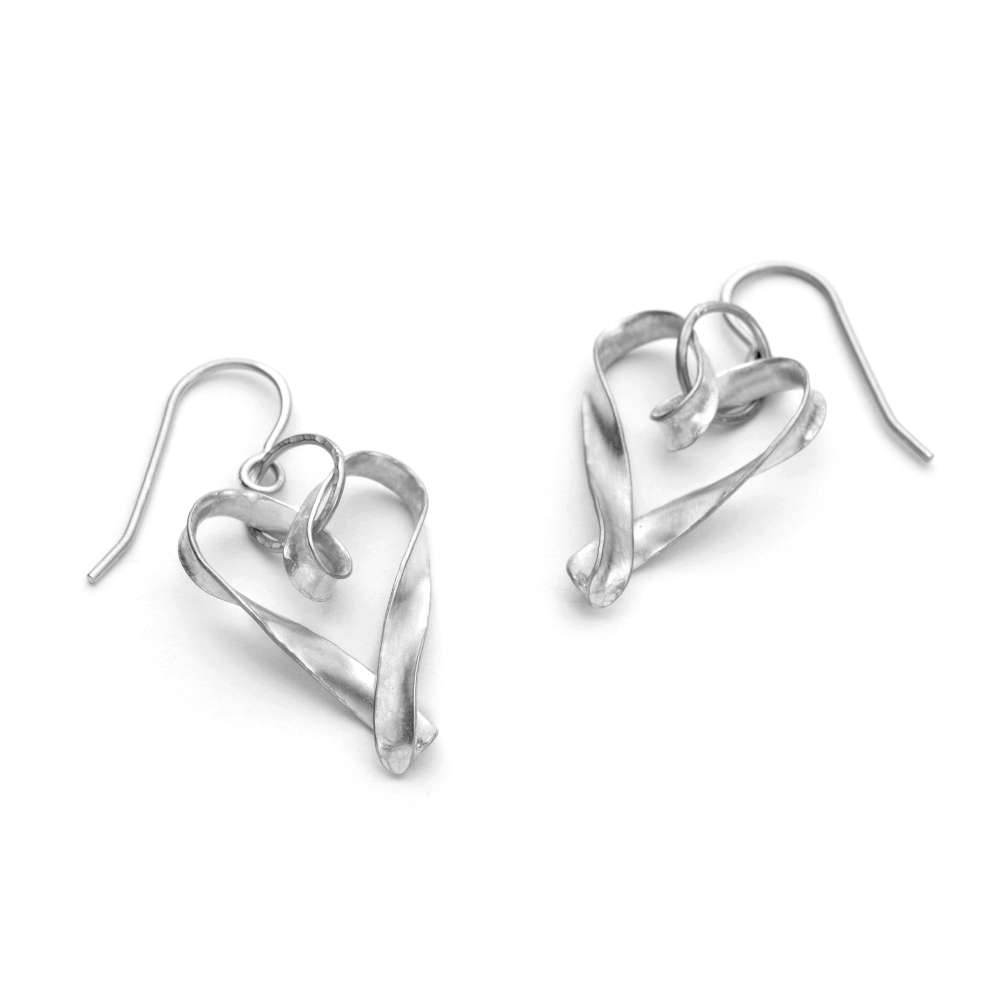 A pair of silver heart drop earrings, each formed from a single ribbon of metal, twisted and joined at the point. They are attached to the ear wire by a large, hammer textured jump ring. They are seen here lying on a surface.