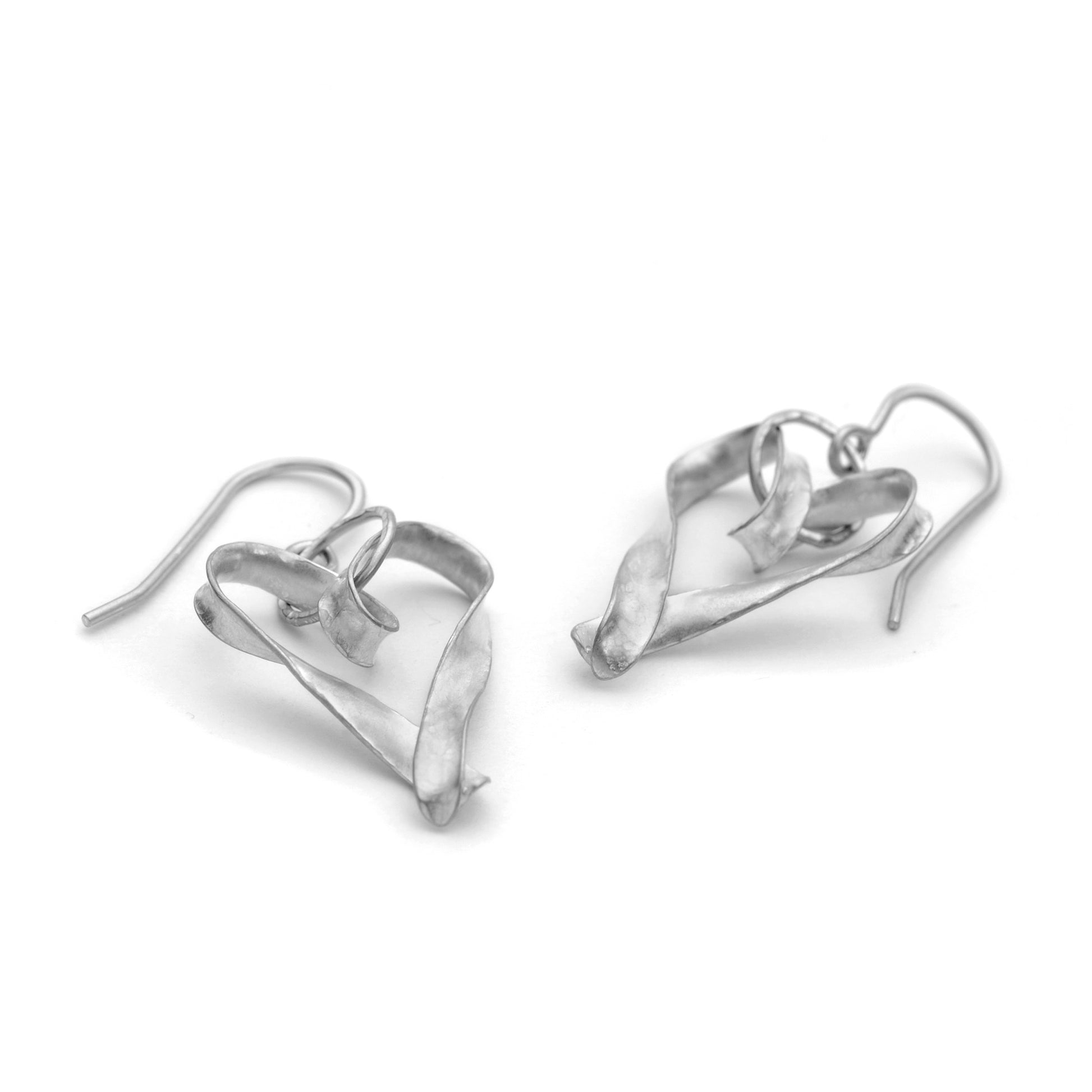 A pair of silver heart drop earrings, each formed from a single metal of silver, curled, twisted and joined at the pointed end. They are attached to the ear wire by a large, hammer textured jump ring. They are seen here lying on a surface and viewed at a low angle from the pointed end.