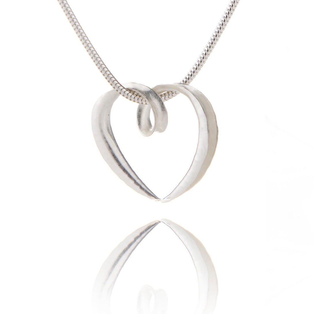 A small silver heart pendant made from a single piece of metal, formed into a sort of tube with the outer side open. It is formed into a loop at the top which the chain passes through.