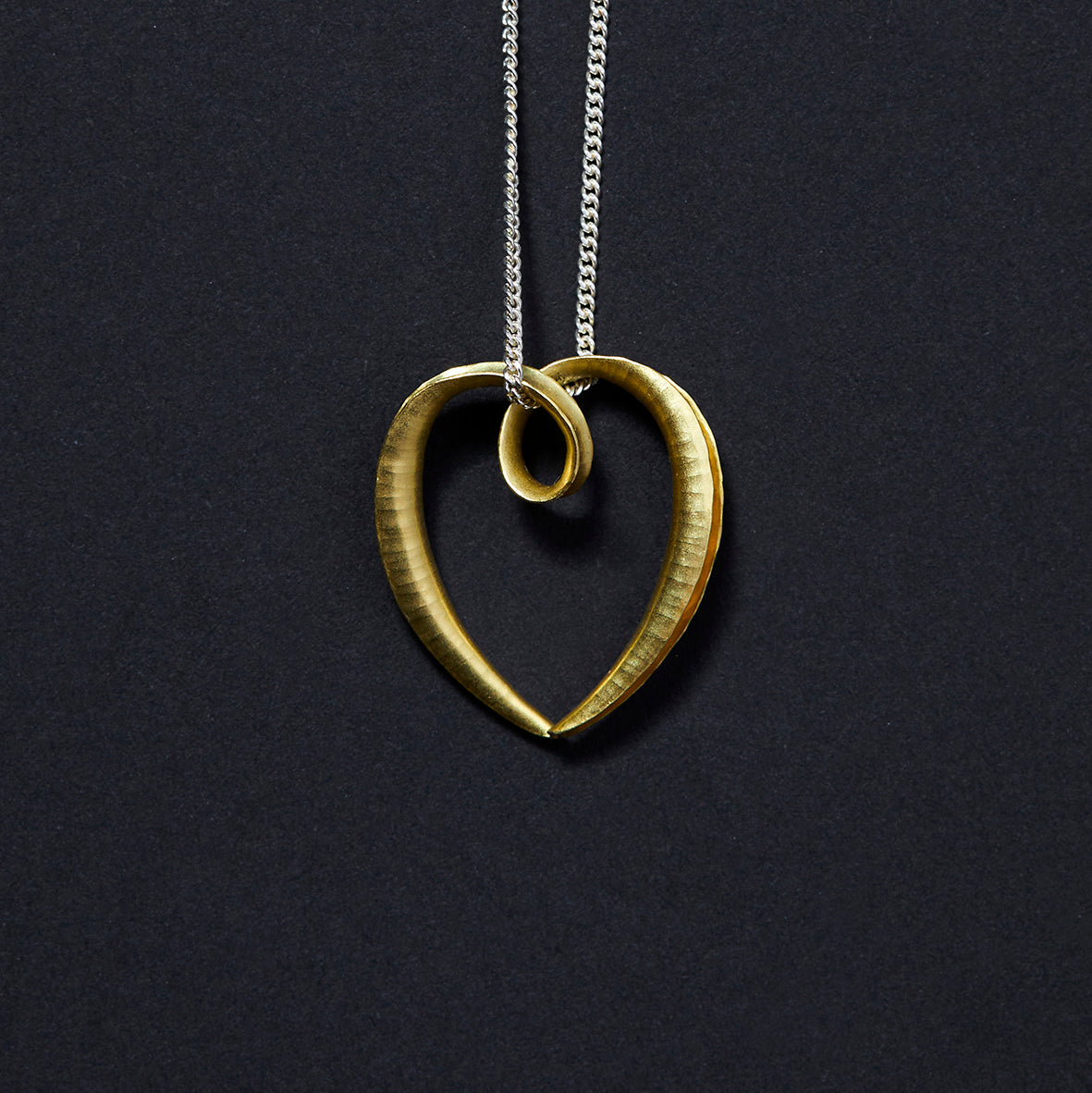 A small 18 carat heart pendant made from a single piece of metal, formed into a sort of tapered tube with the outer side open and gold plated on the inside surface for contrast. It is formed into a loop at the top which the chain passes through.