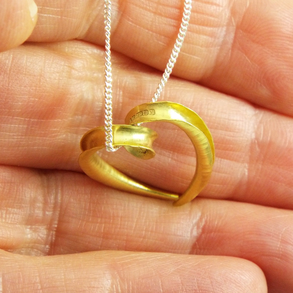 A small 18 carat heart pendant made from a single piece of metal, formed into a sort of tapered tube with the outer side open and gold plated on the inside surface for contrast. It is formed into a loop at the top which the chain passes through. Shown in the hand for scale and to display the hallmark.