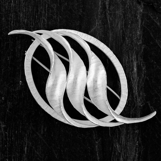 Recycled silver brooch with three repeating twisted s shapes mounted on a narrow oval, with handmade stainless steel double pin, hammered texture and satin-matte finish